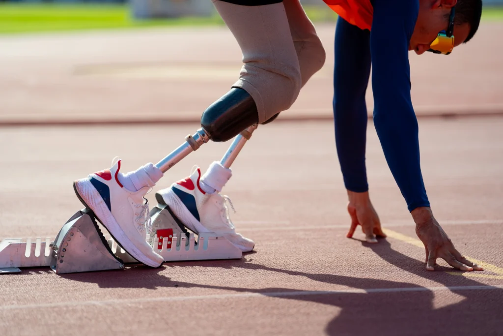 A side profile of a para-athlete with prosthetic legs positioned in metal starting blocks on a running track, ready to begin a sprint.