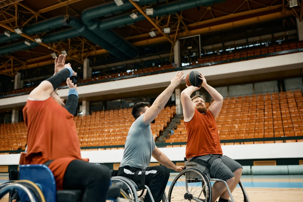 Three athletes playing wheelchair basketball in a large indoor arena; one player in an orange jersey is shooting the ball while a defender reaches to block.