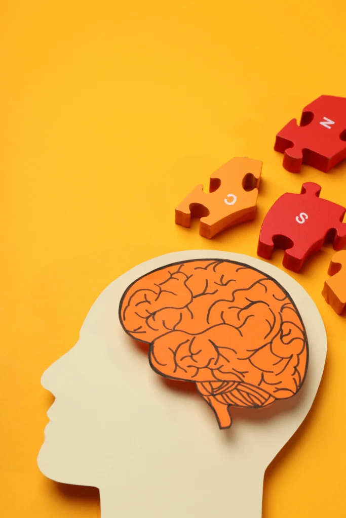 A paper cutout of a human head silhouette with an orange brain illustration inside, surrounded by colorful red and orange puzzle pieces on a bright yellow background.