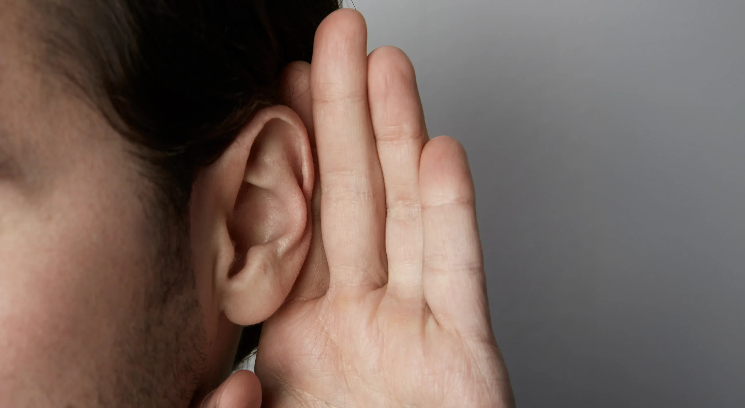A close-up side view of a person cupping their hand behind their ear in a listening gesture against a neutral gray background.