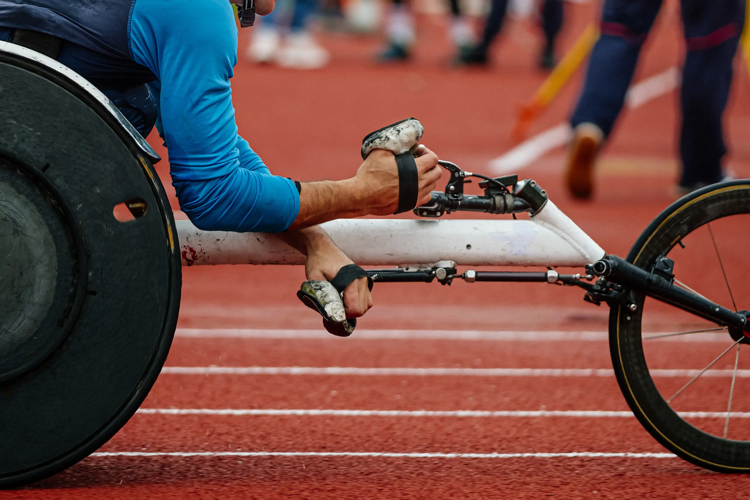 A close-up side view of a Paralympic athlete's hands using specialized gloves to propel a white racing wheelchair on a red running track.
