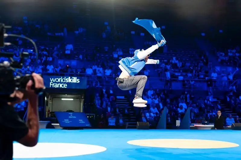 A man in a blue and white varsity jacket performing a mid-air jump while holding a blue flag at a WorldSkills France event in a large arena.