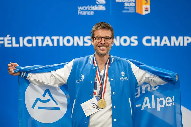 Grégory Cuilleron smiling and wearing a gold medal and blue varsity jacket while holding up a blue flag with a mountain logo at WorldSkills France.