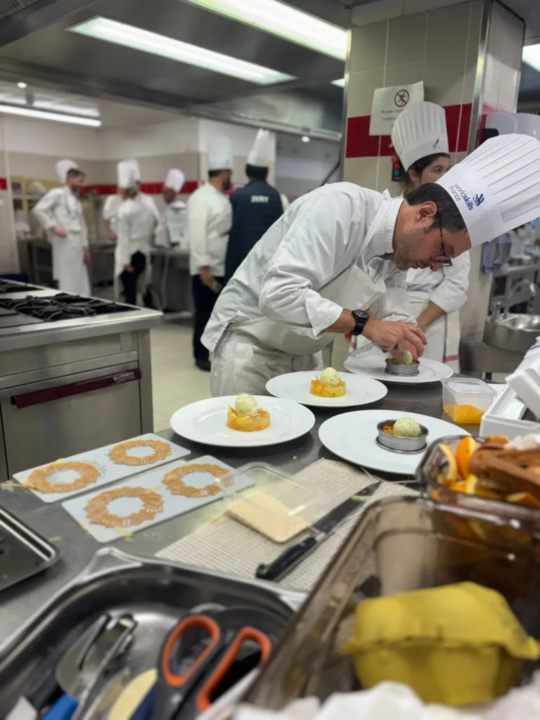 A chef in a white uniform carefully plates food in a professional kitchen during what appears to be a culinary competition or training session. Several prepared plates are arranged on the counter while other chefs work in the background. Stainless steel kitchen equipment and cooking stations surround the workspace.