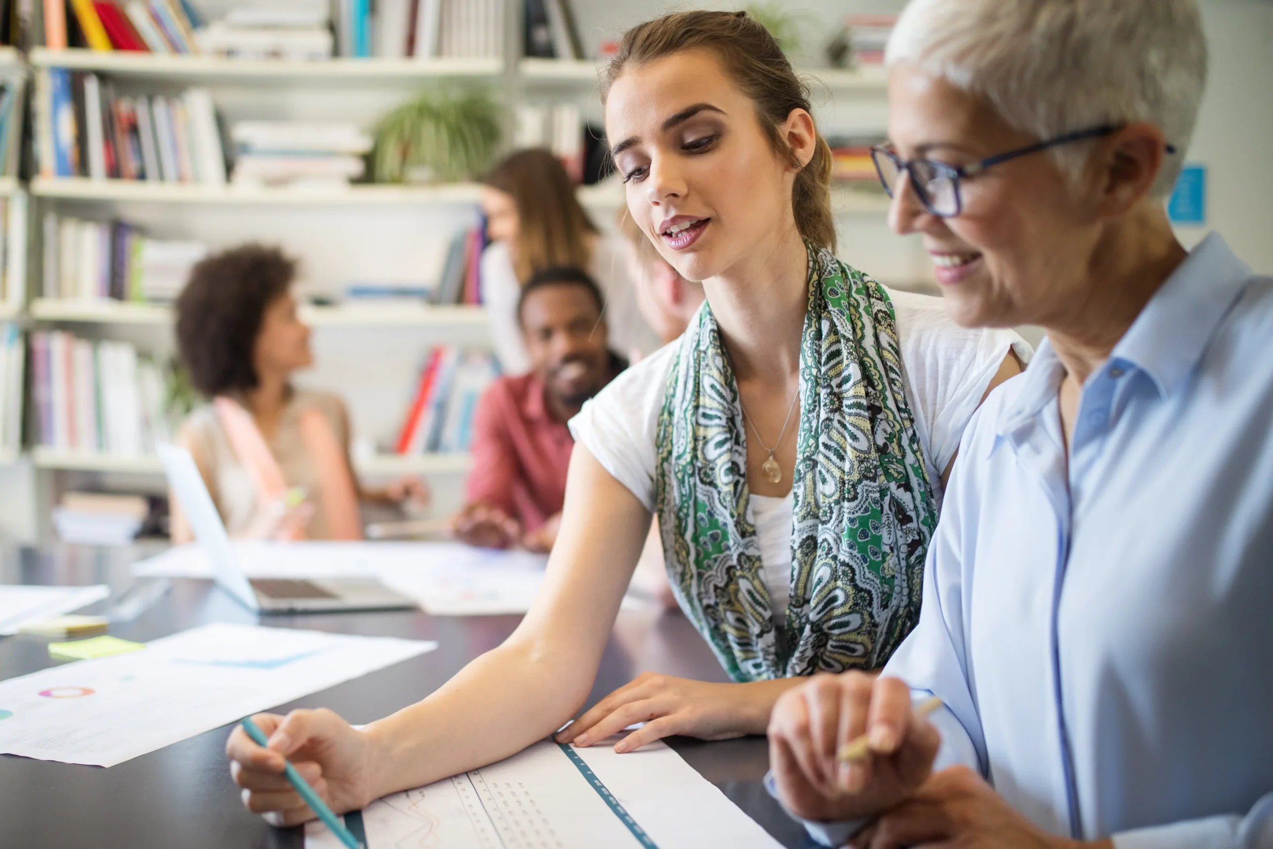 A diverse group of business professionals collaborating in a bright, modern office. A young woman in a green patterned scarf points to a data chart on a desk while discussing it with a smiling senior colleague with short grey hair.