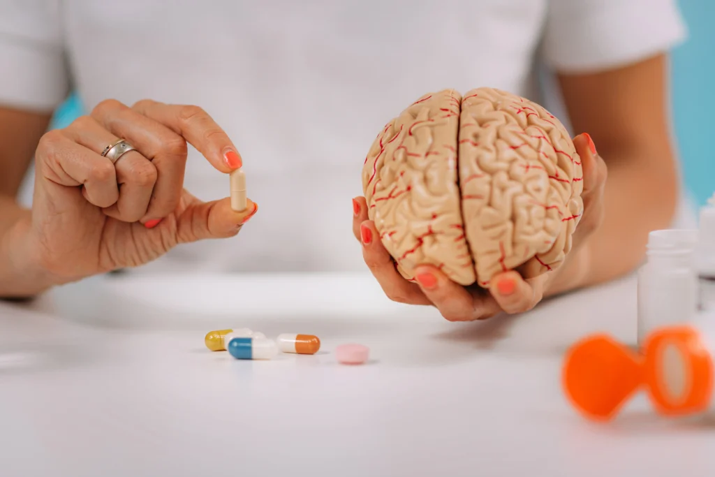 A close-up of a person holding a realistic anatomical model of a human brain in one hand while holding a capsule pill in the other, with various scattered supplements and prescription bottles on a white desk in the foreground.