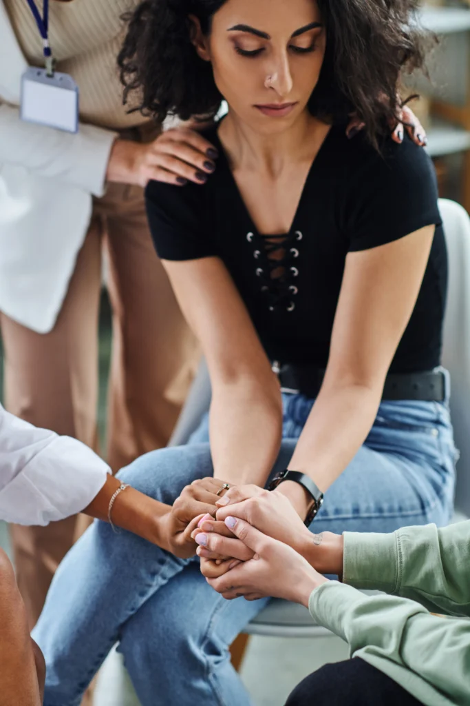 A close-up of a woman with curly hair receiving comfort during a group support session. Several people are shown holding her hands and placing a supportive hand on her shoulder in a gesture of empathy and care.