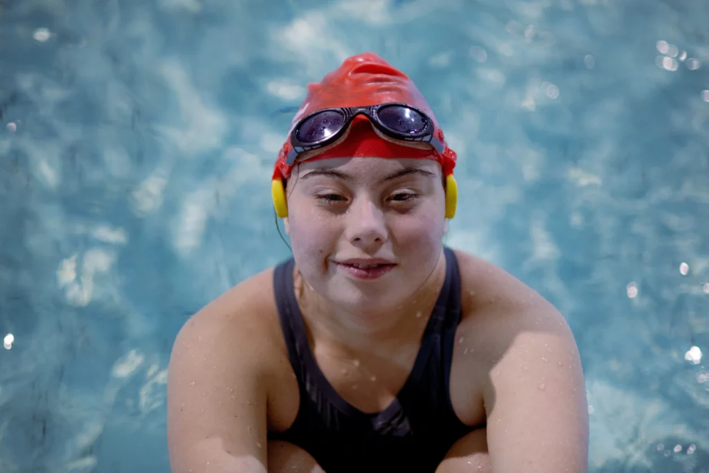 A young woman with Down syndrome wearing a red swim cap and black goggles smiling while treading water in a swimming pool.