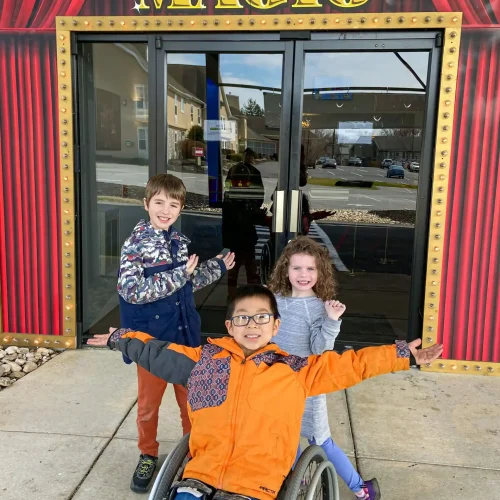 Three children are posing happily in front of a building with a sign that says "MAGIC" in bold, yellow letters. The entrance is decorated to look like a theater, with red curtains and golden accents. One child is in a wheelchair, wearing a bright orange jacket and glasses, extending his arms out in a welcoming gesture. Another child, standing to the left, is smiling widely and gesturing toward the entrance. The third child, standing to the right, is smiling and also pointing toward the entrance. The scene is full of excitement and anticipation.